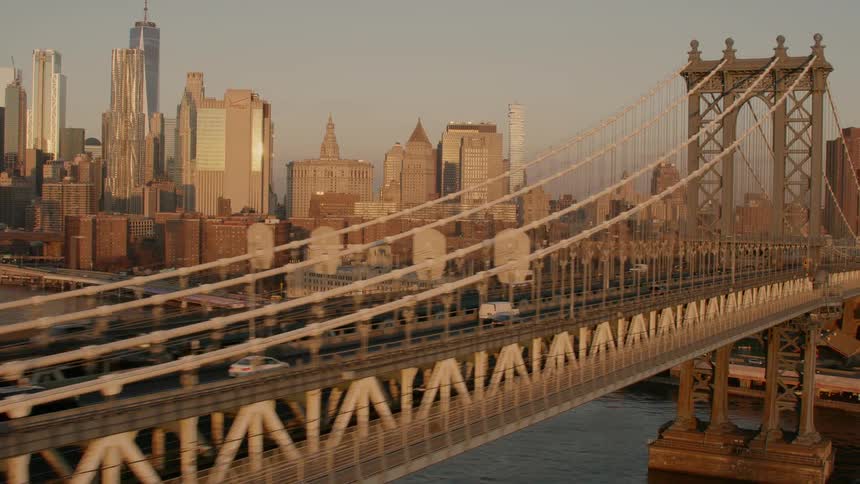 Traffic crosses suspension bridge over water with downtown skyscrapers reflecting sun in the background