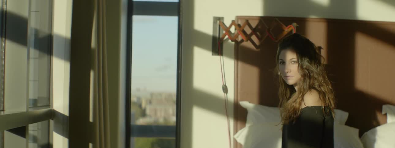 Woman stands by bed looking out the windows