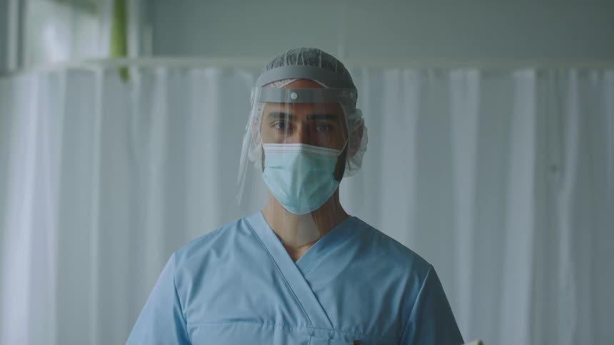 A man with a mask face shield and scrubs in the hospital room with a wall and window in the background