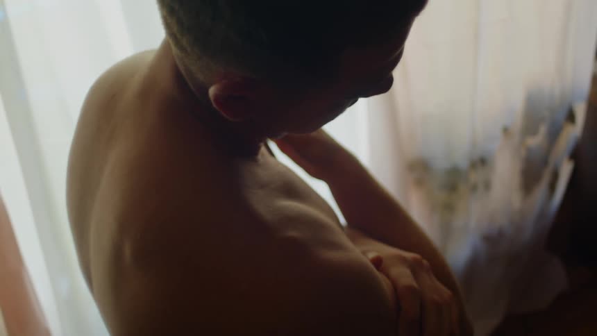 Man stands bare chested in living room stretching and rubbing neck and shoulder