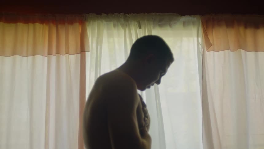 A shirtless man is standing while rubbing his upper body and violently convulsing while in a home during the day
