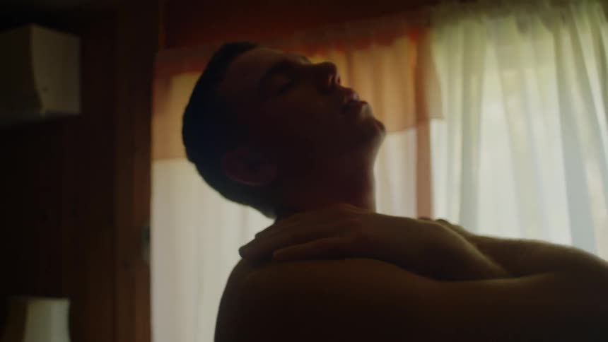 A man is standing in a room in front of the window rubbing his chest and shoulders