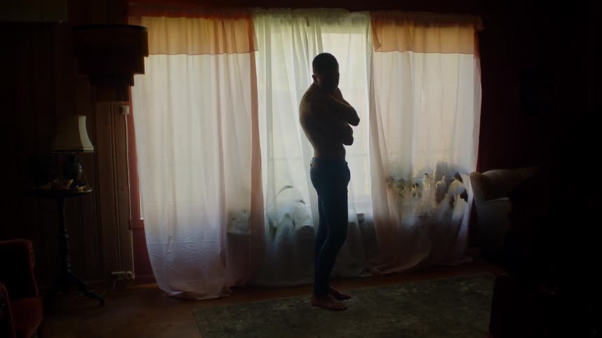 Shirtless man with pants stands next to window