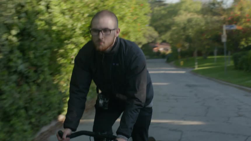 A man with glasses and a camera riding a bicycle in a neighborhood street with the sky and clouds in the background