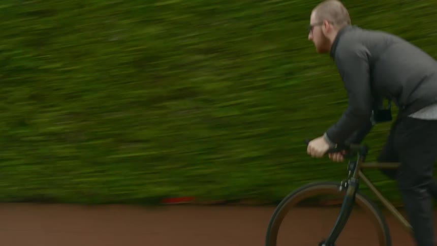 Man with glasses beard black coat and pants pedals along the street fast