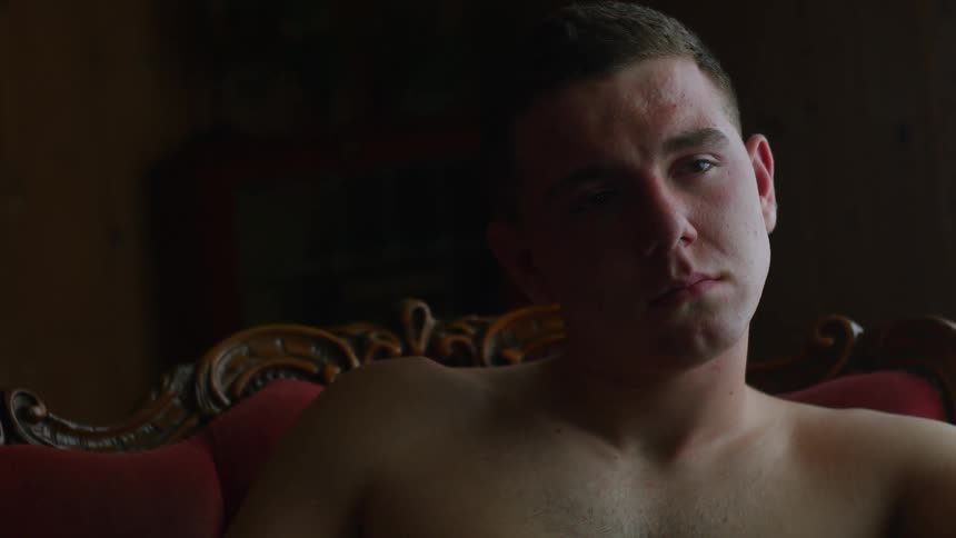 A bare chested man is sitting in a chair in the darkness