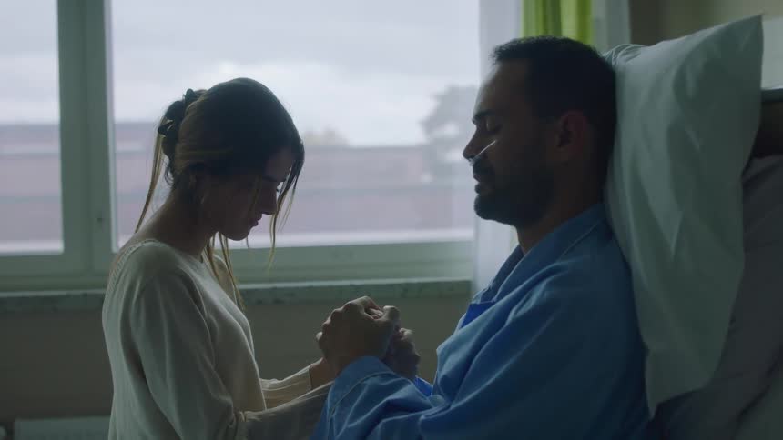 A woman is sitting and holding hands with a man in a hospital bed while they both pray with their eyes closed in the daytime