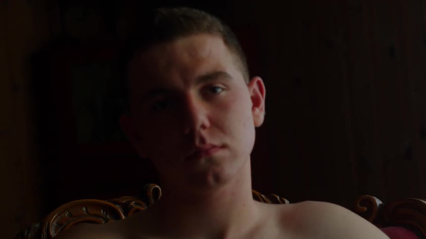 A shirtless man sits in a chair with his head cocked to the side