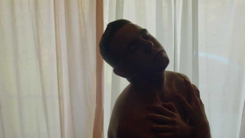 Young shirtless man rubbing his chest in front curtains at home