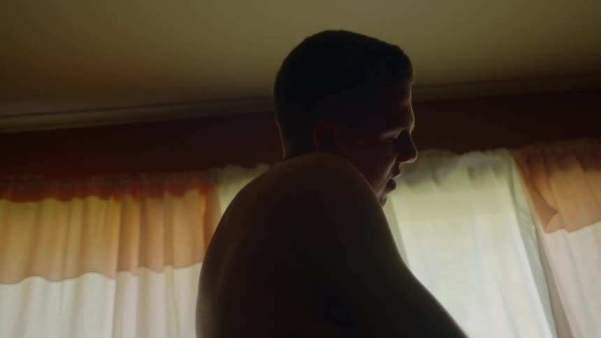Young shirtless man rubbing his arms and chest inside a dimly lit room at home