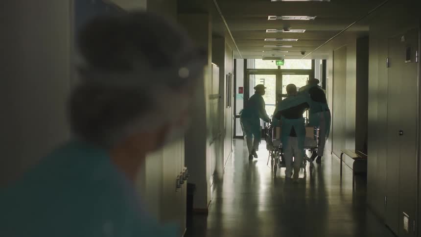 Medical personnel wheel a patient in a stretcher down a hall as a nurse watches and then rushes to catch up