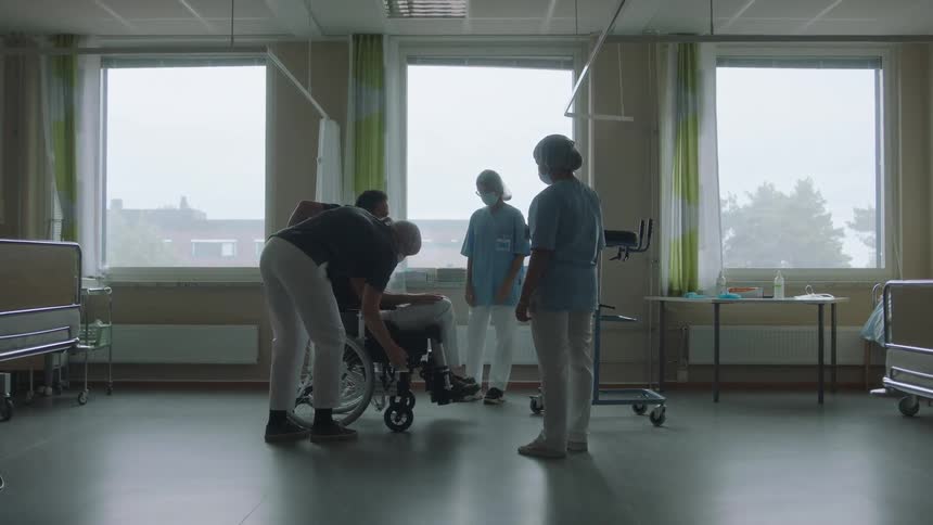 A man in a wheelchair in the hospital room with three nurses with a wall and window in the background
