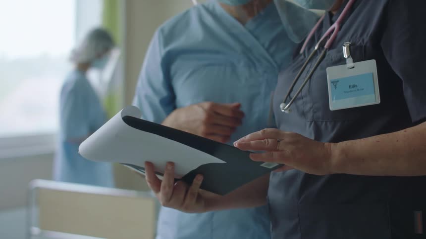 Nurse listens to more senior medical personnel discuss patient chart as they stand in hospital room