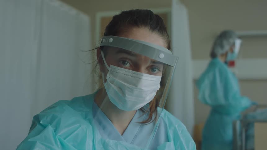 Young female nurse wearing face shield and mask working at the hospital looks at the camera