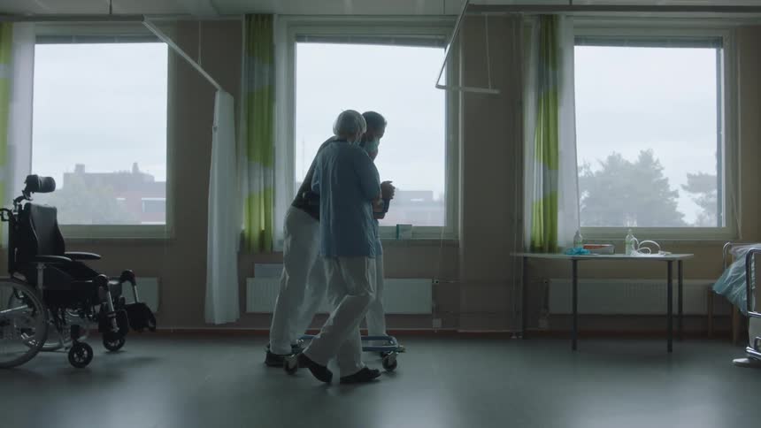A man leaning on a walker with a nurse on either side takes steps away from a wheelchair