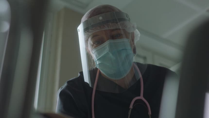 Female nurse wearing face shield and mask talking to a patient on a hospital bed