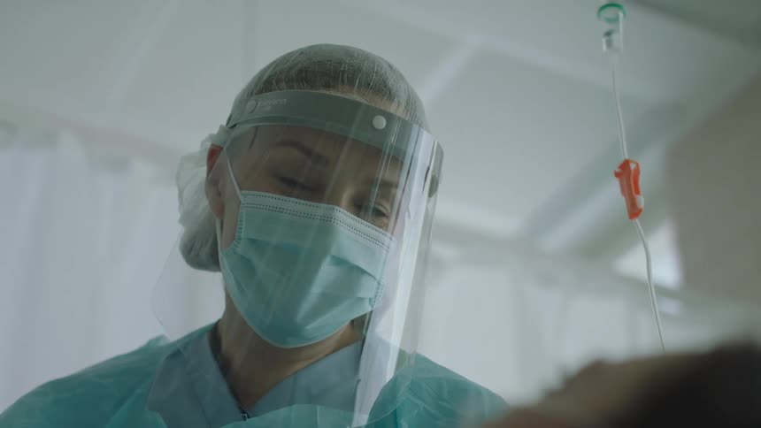 Nurse wearing face shield and face mask talks to patient lying down with iv line