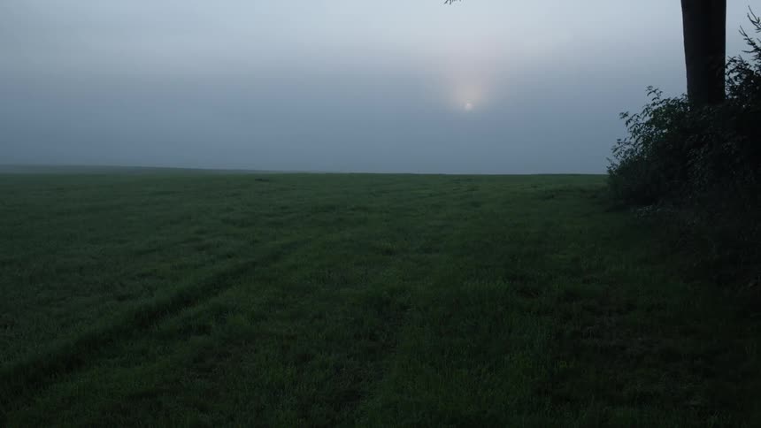 Sun shines dimly through clouds onto mowed field bordered by brush