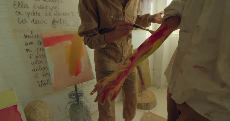 A woman in a boy in a room with the woman painting on the boys arm with a wall and window in the background