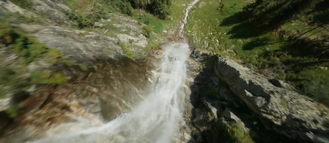A waterfall leads to a small river at the bottom of a mountain range surrounded by trees