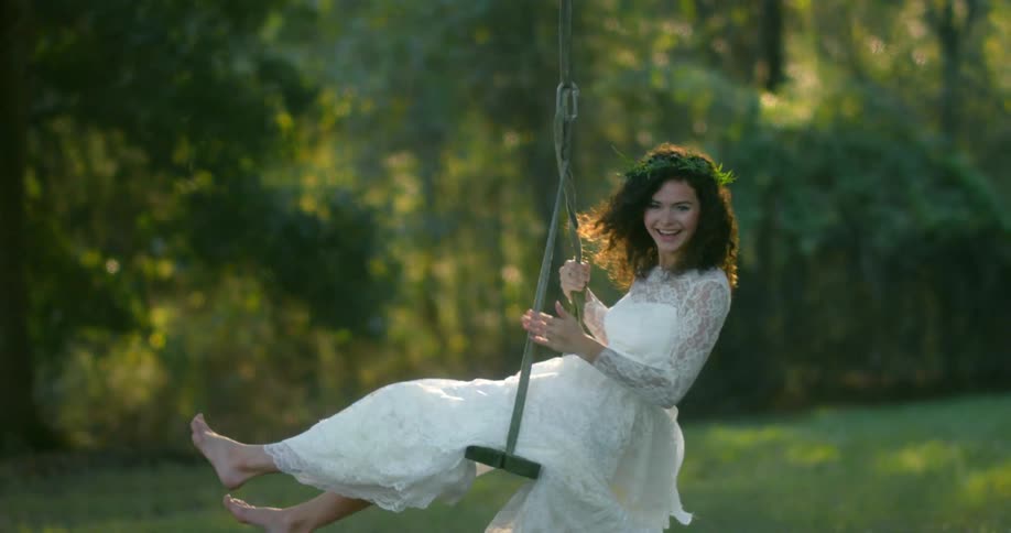 A woman in a wedding dress is smiling and laughing as she swings on a wooden rope swing