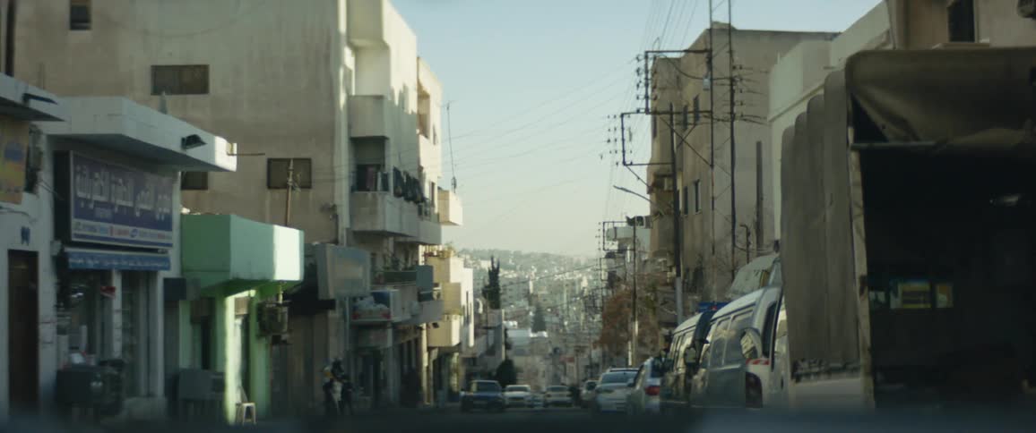 People stand near storefront across road from parked vehicles with town stretching from base of hill to horizon