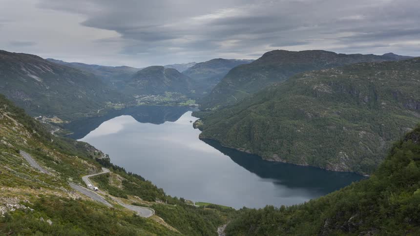 Cars travel on a road near a lake between mountains under a cloudy sky