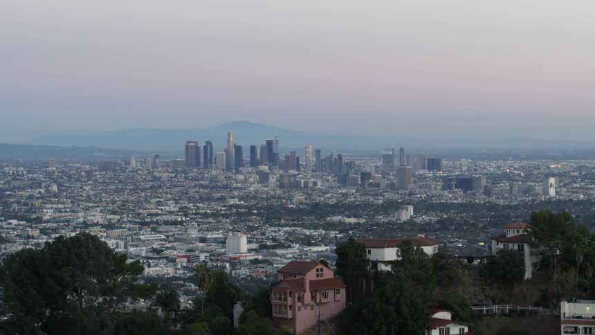 Mansions in Hollywood Hills frame the DTLA skyline and mountains covered in dense haze under a pink tinted sky