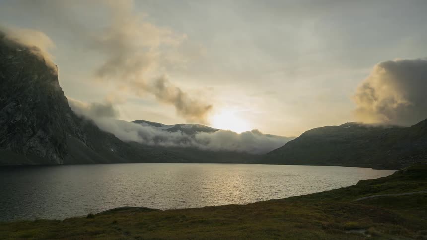Clouds move over a lake surrounded by mountains as the sun shines brightly on the horizon