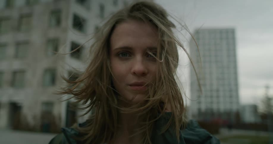 Blonde woman stands with building in background as wind blows her blonde hair about