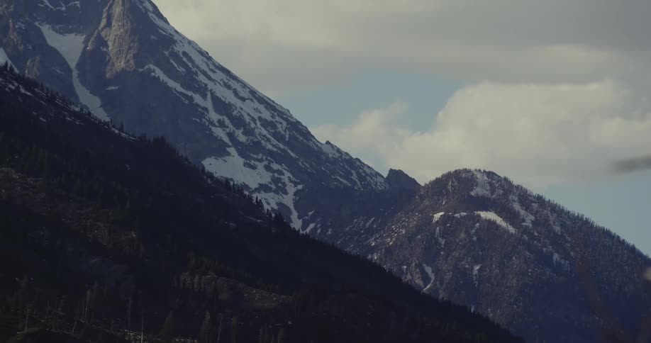 A mountain range with trees and snow with the sky and clouds in the background