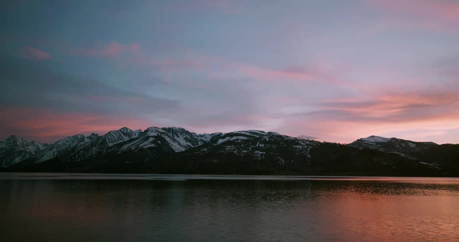View of still lake with snowy mountain ranges under red sky at dusk