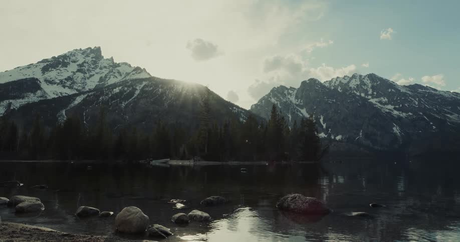 Water moves along the rock covered shore as the sun rises over snow covered mountains