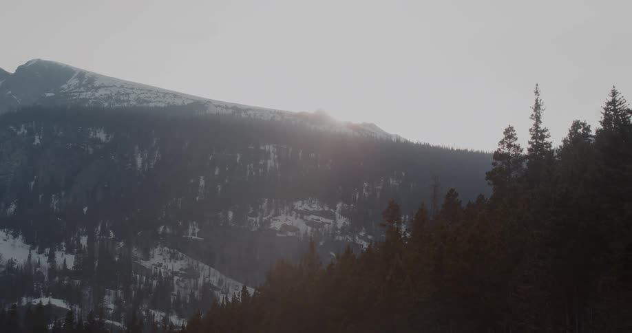 Sun is setting over mountains partially covered in snow and trees