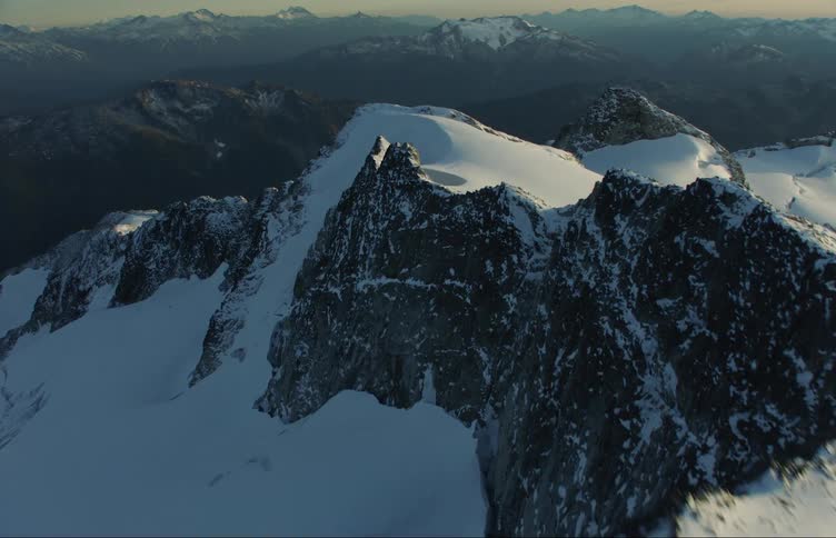 Aerial view moving across the top of a snow covered mountain ridge and a mountain range on the horizon