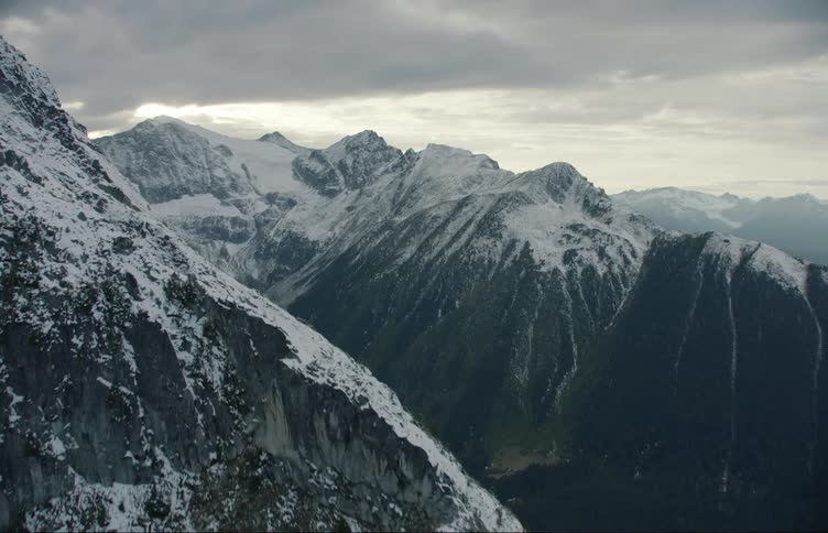 Snow covers mountains and forested valley under cloudy sky