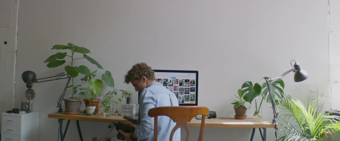 Photographer sits at table reviewing photographs on monitor while holding camera