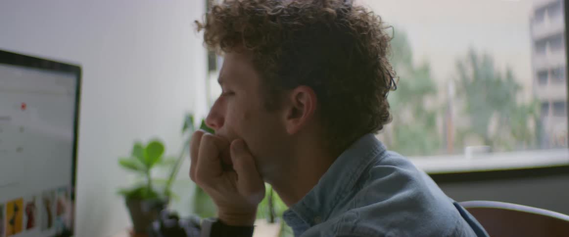 A man sits at a desk with a camera and looks at photos on his computer