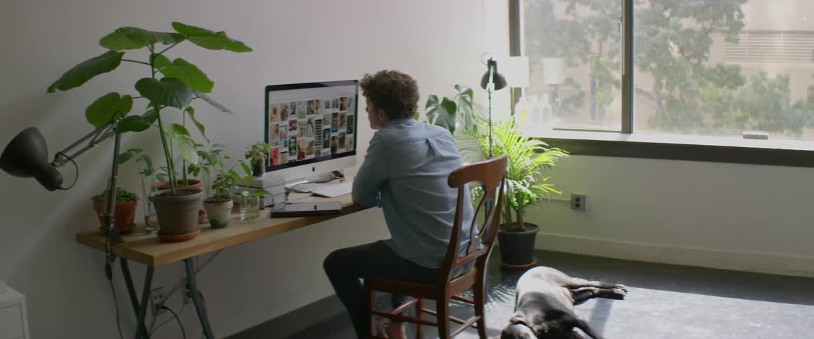 A man sits at a desk at home and looks at photos on his computer as his dog lies behind his chair