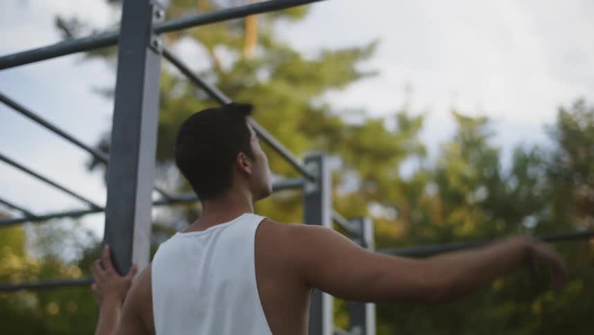 Man wearing tank top walks up to metal structure and reaches up to grasp metal bar under cloudy sky
