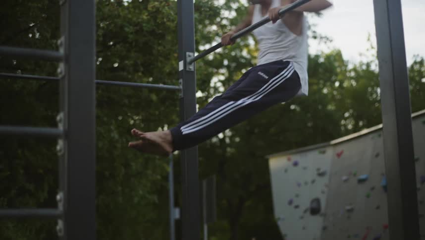 Man wearing tank top exercises on metal bar as another person climbs on climbing wall in the background