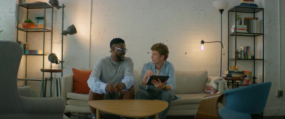Two men in a room sitting on a couch looking at a tablet with a wall in the background