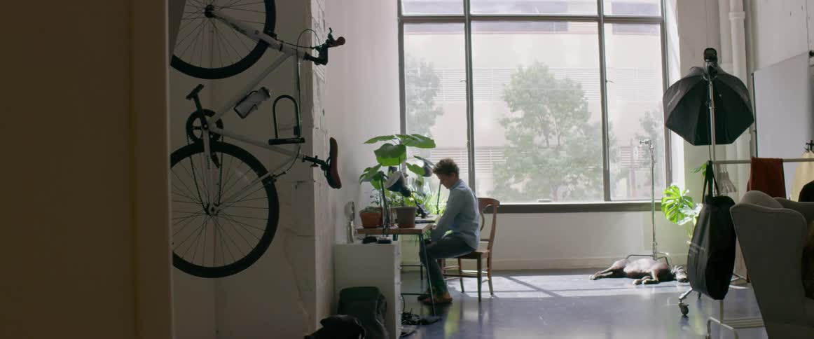 A man walking in a room and sitting on a chair at a table with a wall and window in the background