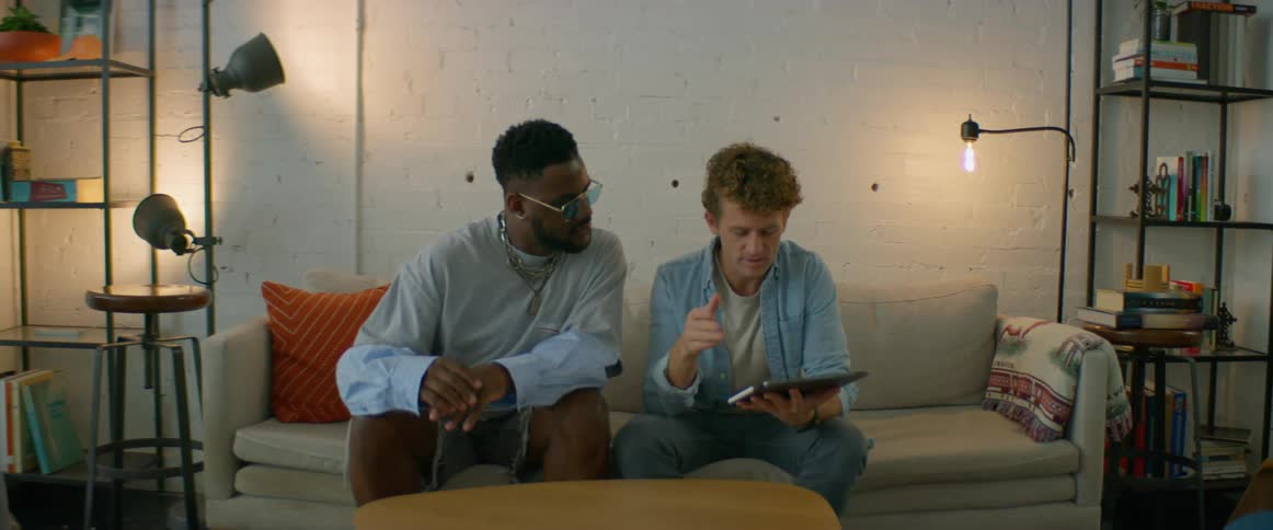 Two men walk over to a couch and sit to discuss items shown on a tablet