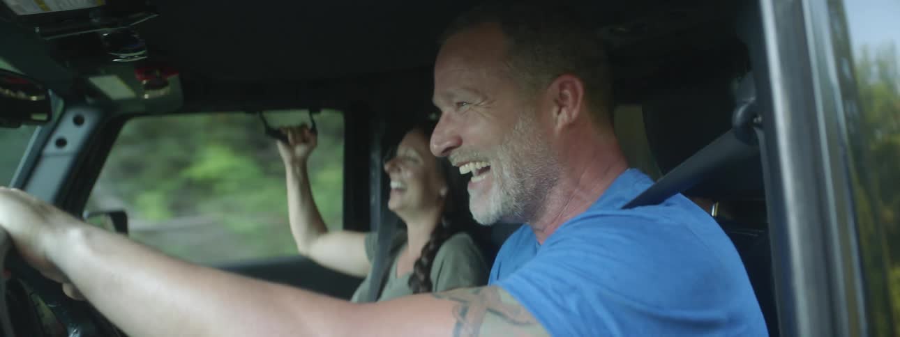 A man pumps his fist in the air as he drives and talks and laughs with the woman in his vehicle