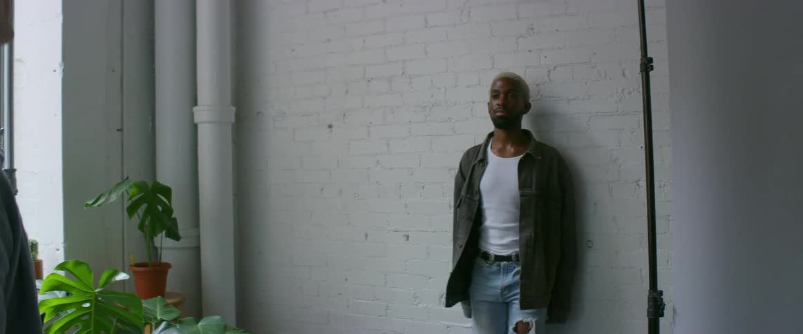 A photographer is taking pictures of a black man posing against a wall