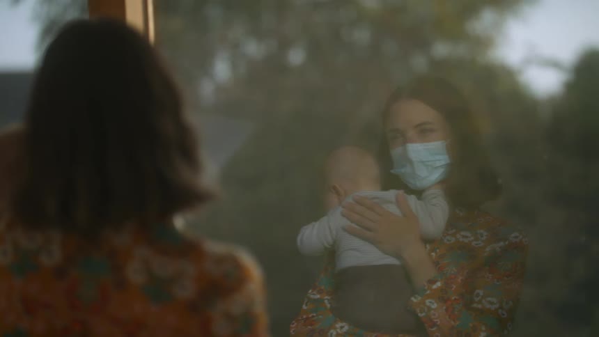 Woman wearing face mask looks out window that reflects her image as she rubs babys back