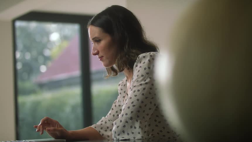 A woman in a room sitting at a table using a laptop with a wall in the background