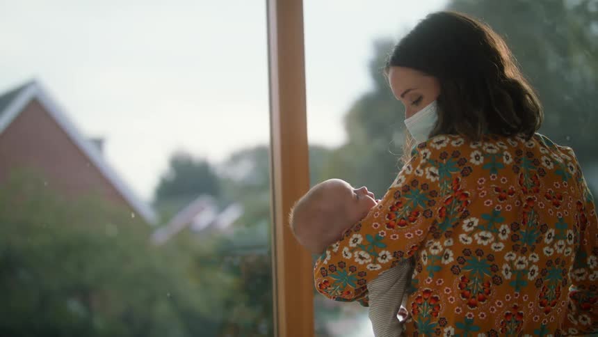 Mother with wavy dark hair mask and orange blouse with flowers swings baby to sleep in front of windows