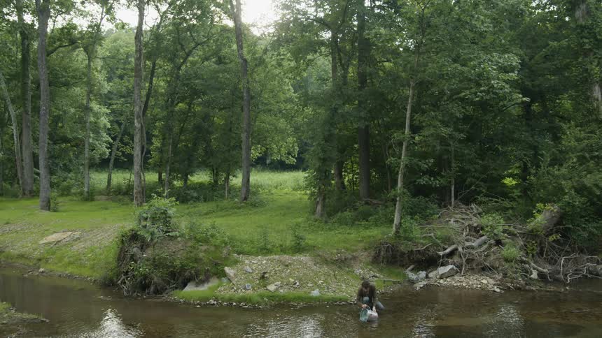 A man dipping a bucket into a river with the trees the sky and clouds in the background